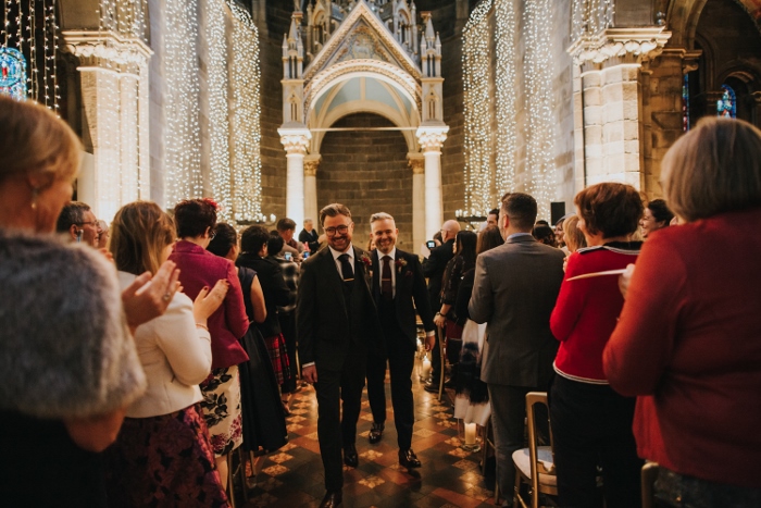 A newly married couple walking back down the aisle, married, to a backdrop of  family, friends and fairylights